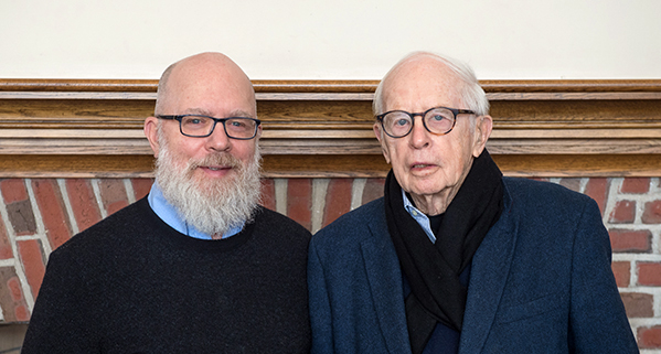 Two men wearing glasses pose in front of a fireplace